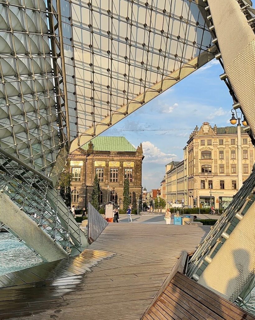 Modern mesh structure framing a wooden boardwalk with a view of historic buildings and people strolling.