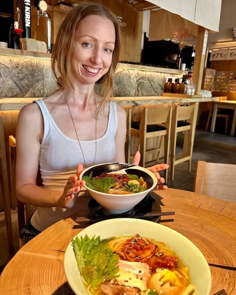 Smiling woman in a white tank top sits at a wooden table holding a bowl of ramen.