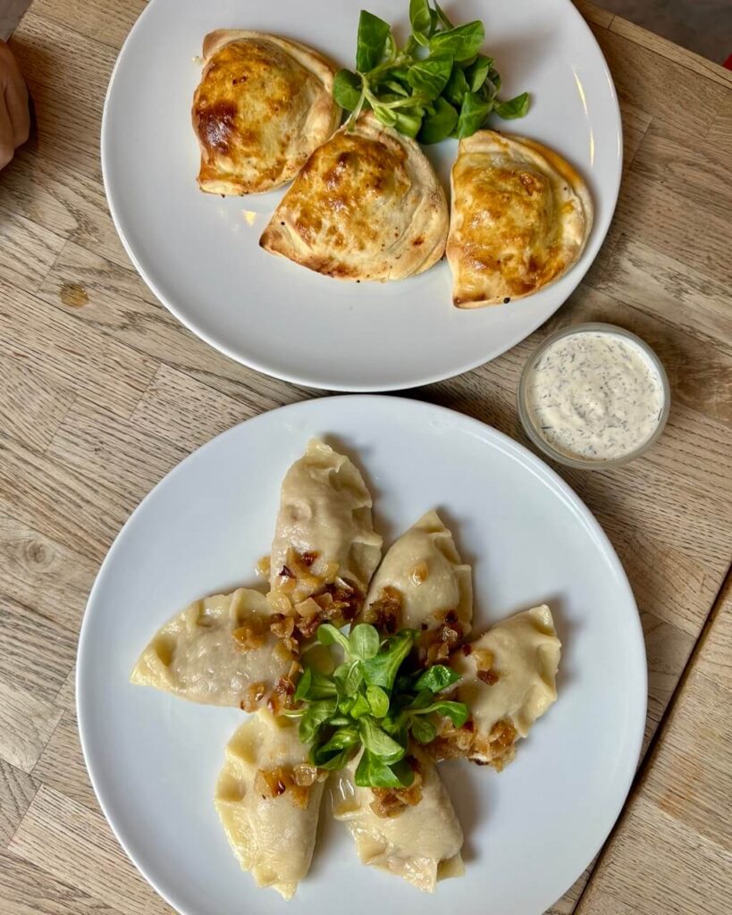 Two plates of pierogi, one baked and one boiled, garnished with greens and fried onions, with dill sauce on the side.