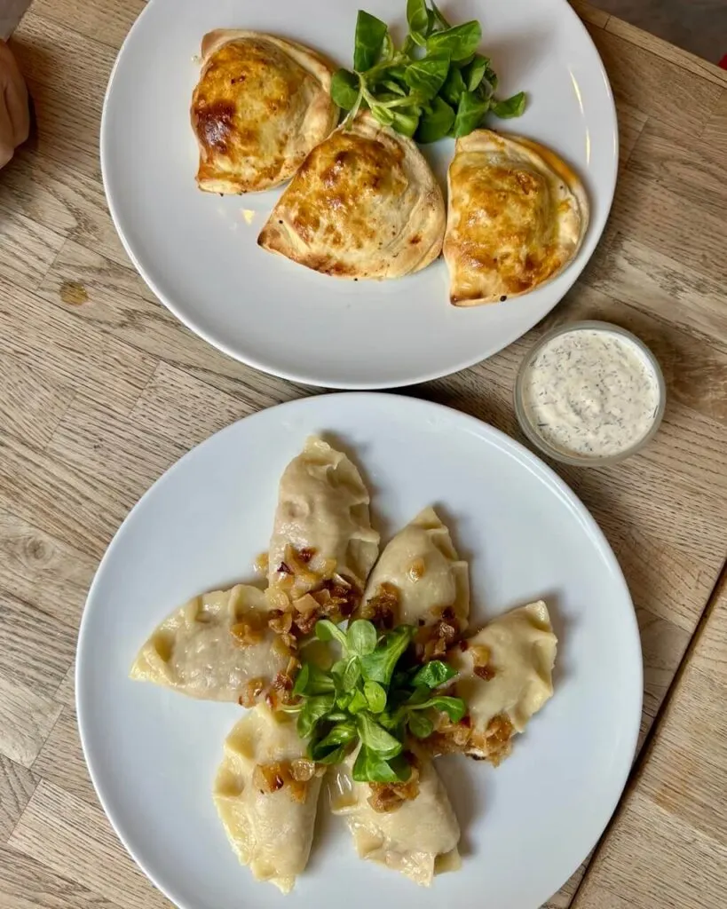 Two plates of pierogi, one baked and one boiled, garnished with greens and fried onions, with dill sauce on the side.