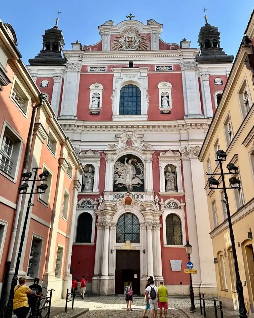 View of the pink and white Baroque facade of the Fara Poznańska church framed by narrow Old Town streets.