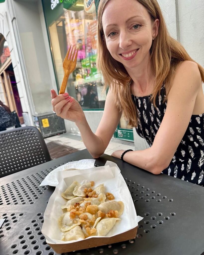 Woman smiles while sitting at an outdoor table with a tray of boiled pierogi topped with crispy bacon bits.