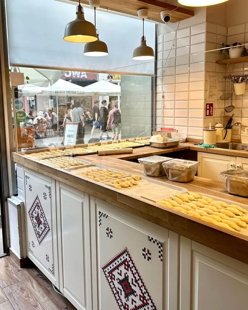 Rows of handmade dumplings are laid out on a wooden counter inside a kitchen with a street view window.