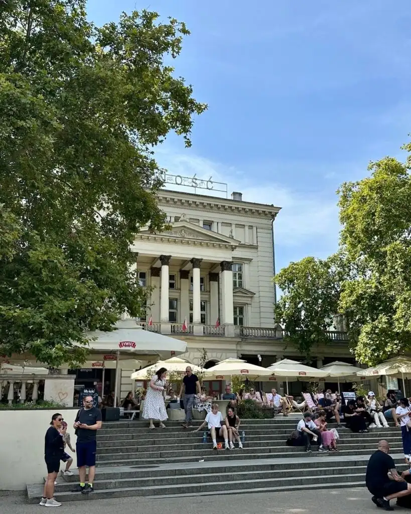 People sit and gather on steps and at outdoor café tables in front of a neoclassical building with columns.