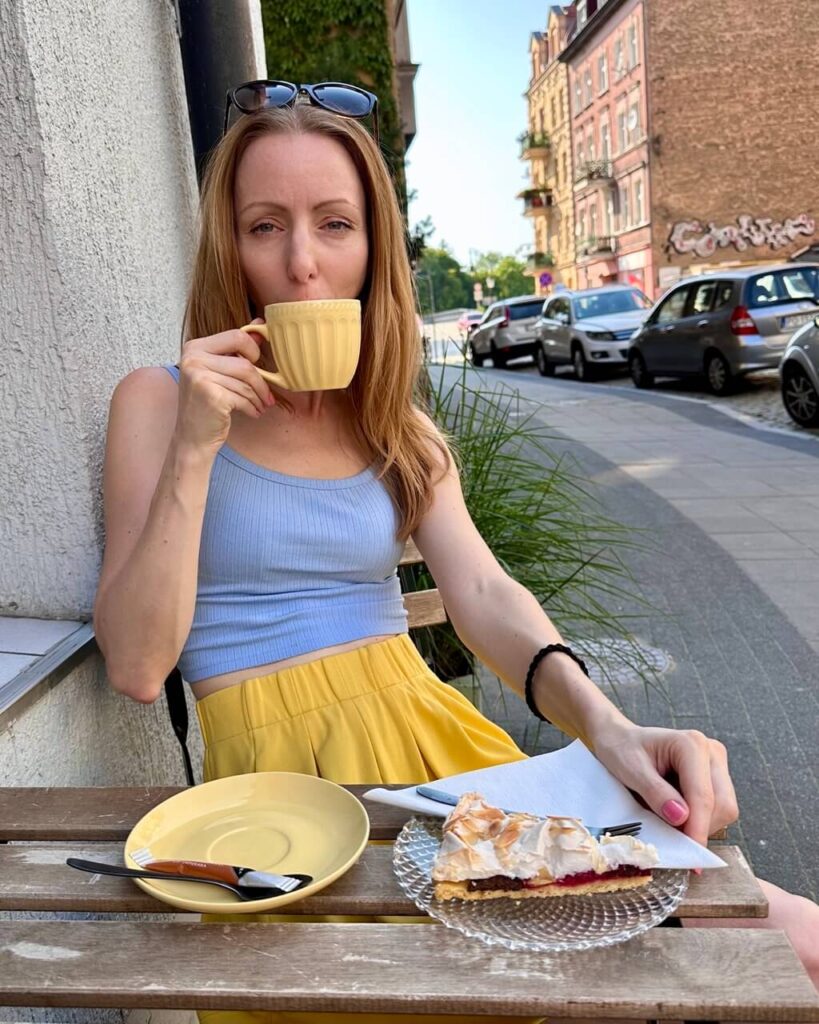 Woman in a blue top and yellow skirt enjoys coffee and a slice of cake while sitting at a sidewalk café.