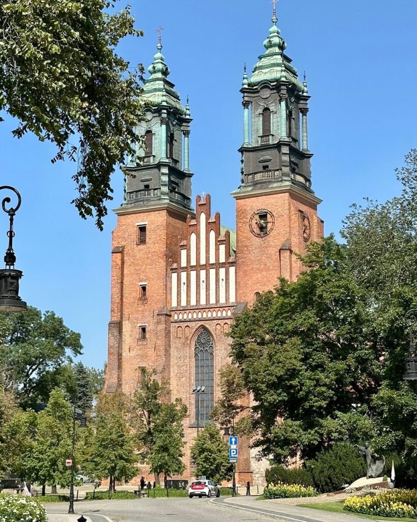Brick twin-towered cathedral with green spires and Gothic windows, framed by trees.