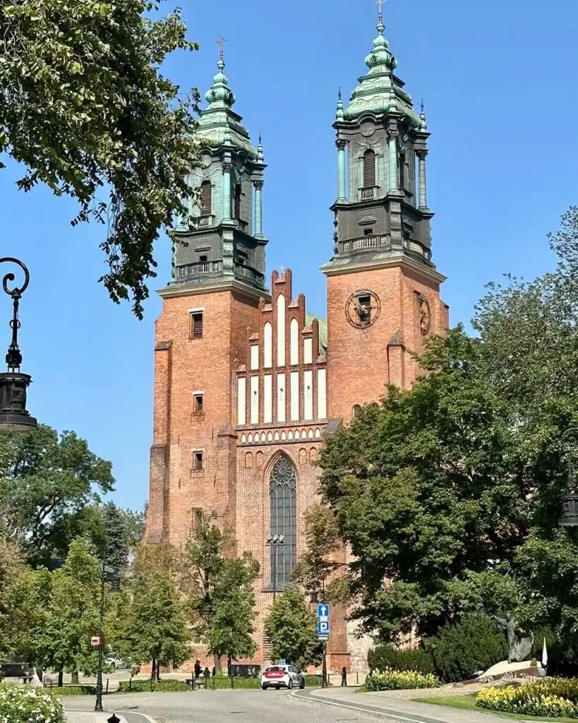 Brick twin-towered cathedral with green spires and Gothic windows, framed by trees.