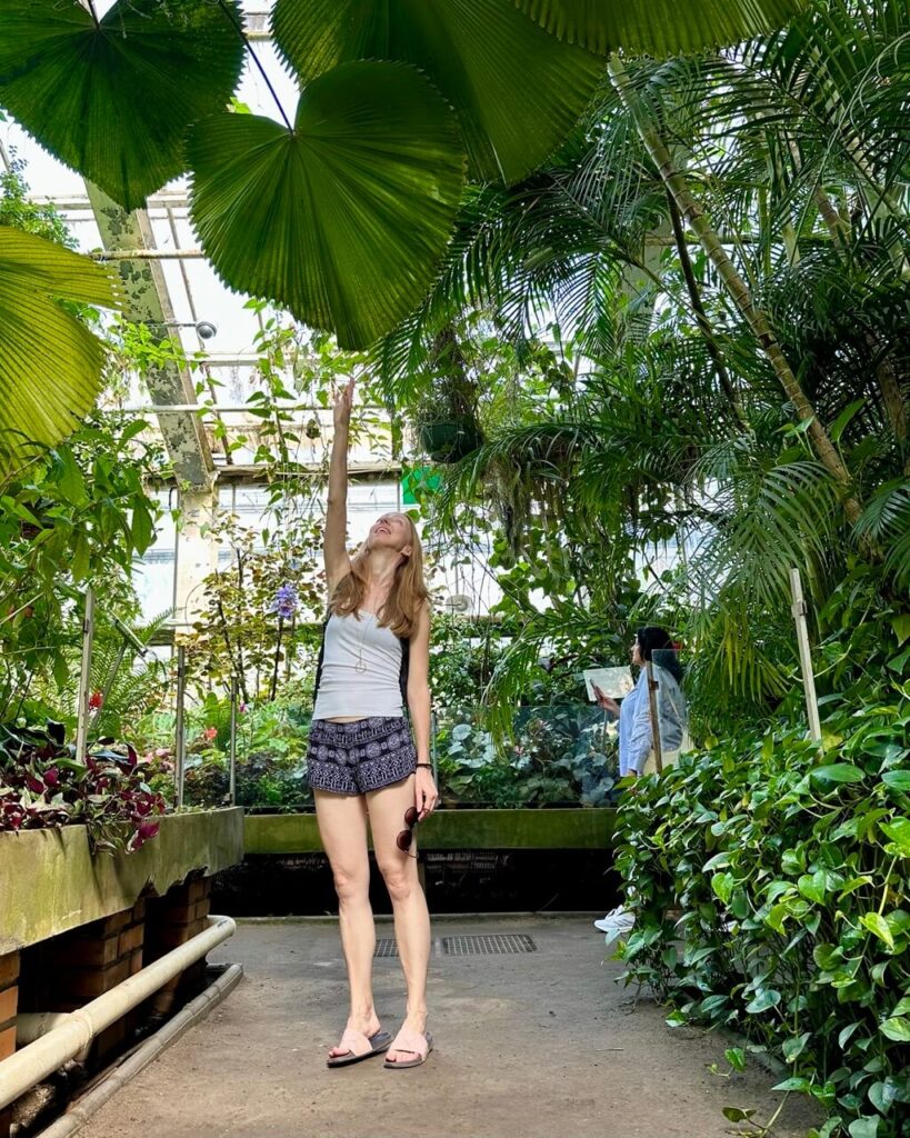 Woman reaches toward a massive heart-shaped tropical leaf inside a lush greenhouse.