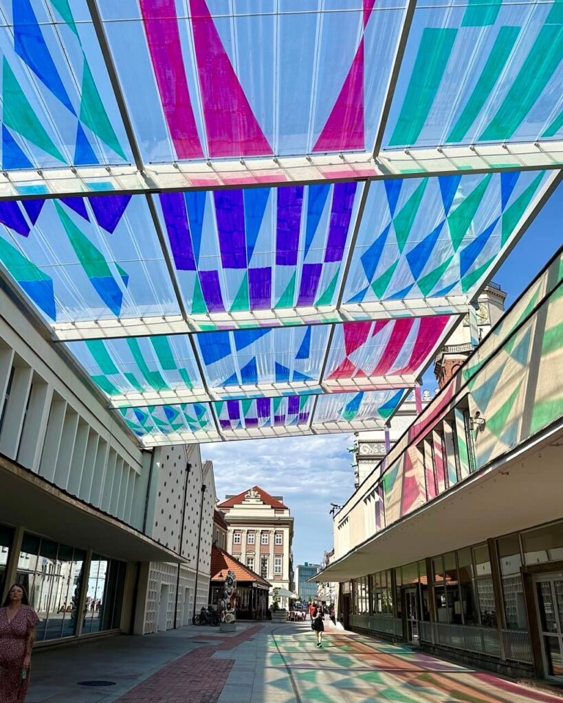 Street lined with modernist buildings is covered by a colorful geometric fabric canopy that casts shadows on the ground.