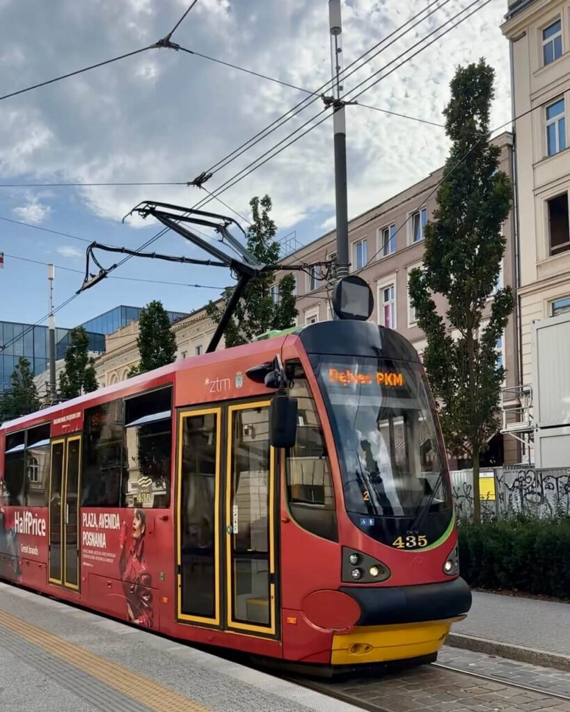 Red tram with yellow accents travels under overhead cables along a city street in Poznan Poland.