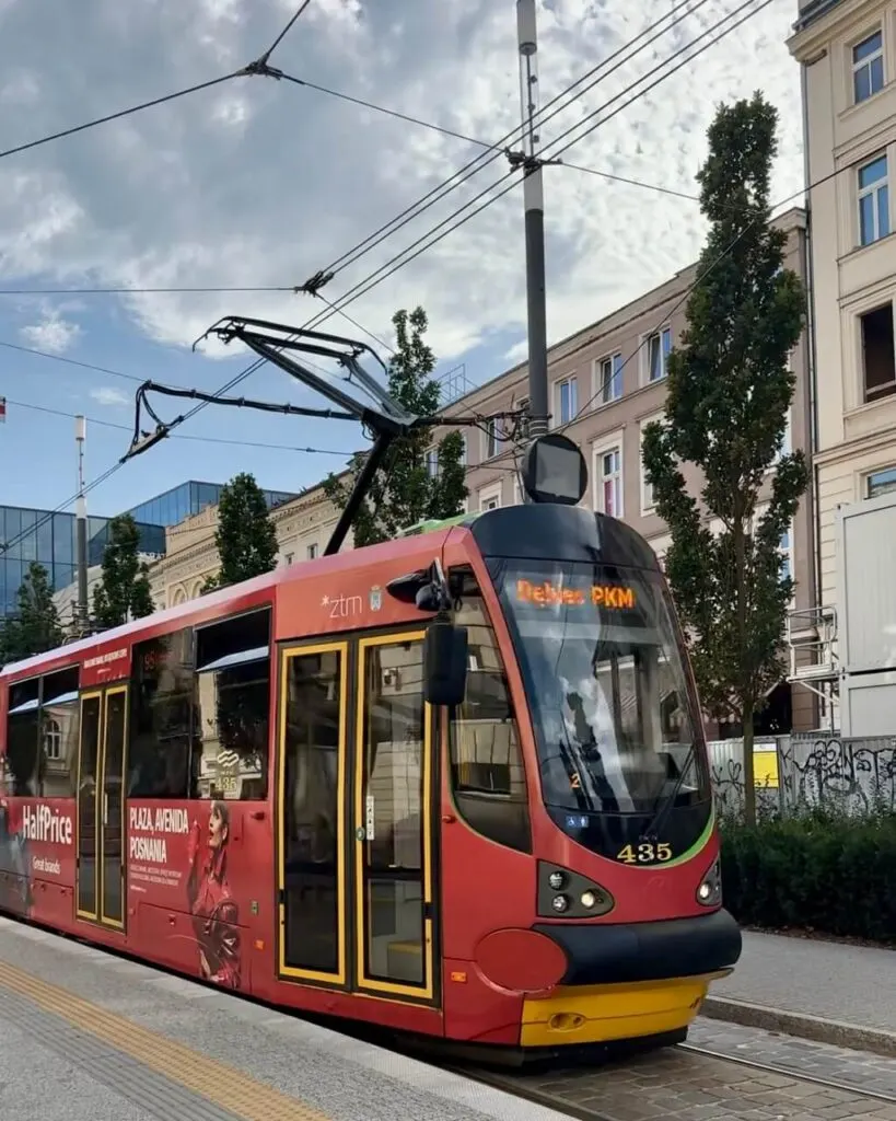 Red tram with yellow accents travels under overhead cables along a city street in Poznan Poland.