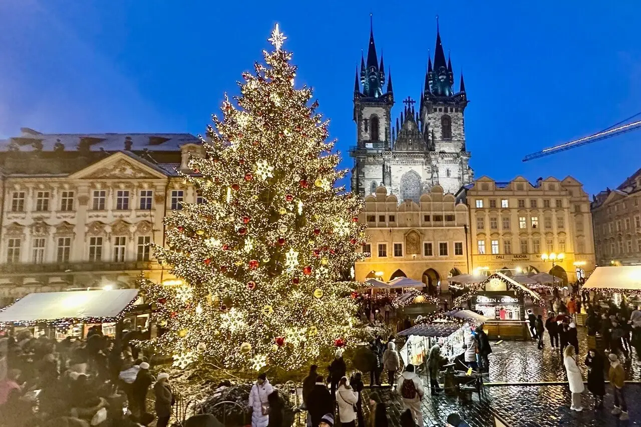 A brightly lit Christmas tree stands in the center of Prague’s Old Town Square, surrounded by festive market stalls and a crowd of people, with the Church of Our Lady before Týn illuminated in the background.