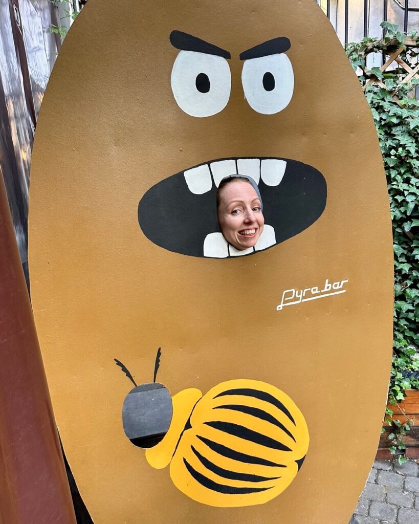 Woman poses with her face through a cutout in a cartoon potato board painted with teeth and a striped beetle at Pyra Bar.