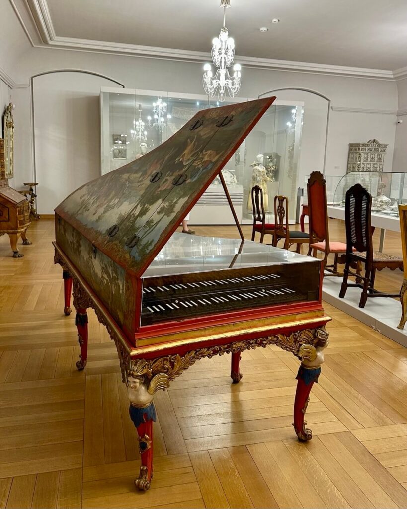 Ornate harpsichord with painted lid and gold-trimmed legs on display in a museum room with antique furniture.