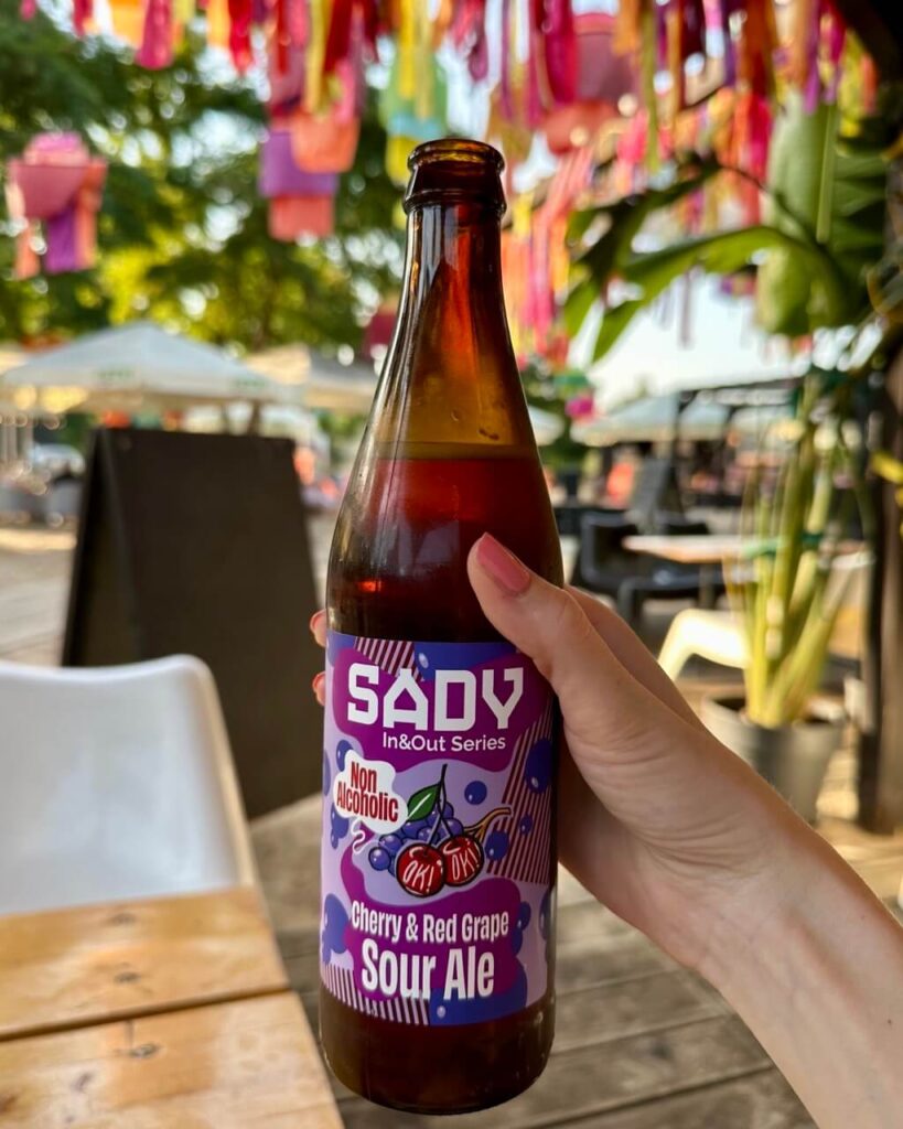 Hand holds a bottle of Sady cherry and red grape sour ale in a colorful outdoor bar decorated with lanterns.