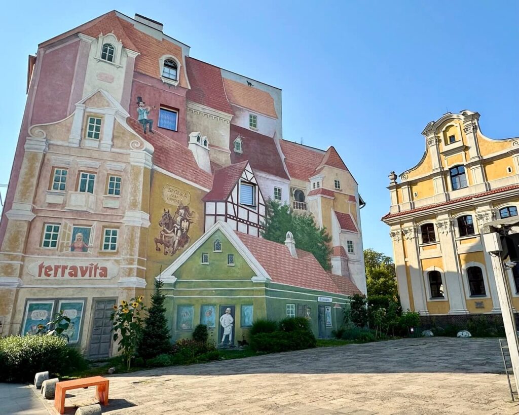 Building façade painted with 3D-style murals of houses, windows, and figures including a man on a roof and a knight on horseback.