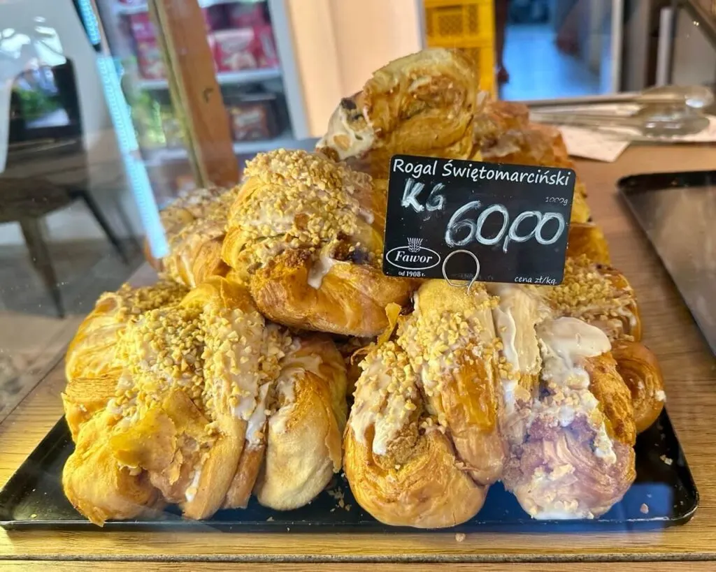 Glazed croissants topped with chopped nuts sit under a glass counter with a handwritten sign reading “Rogal Świętomarciński.”