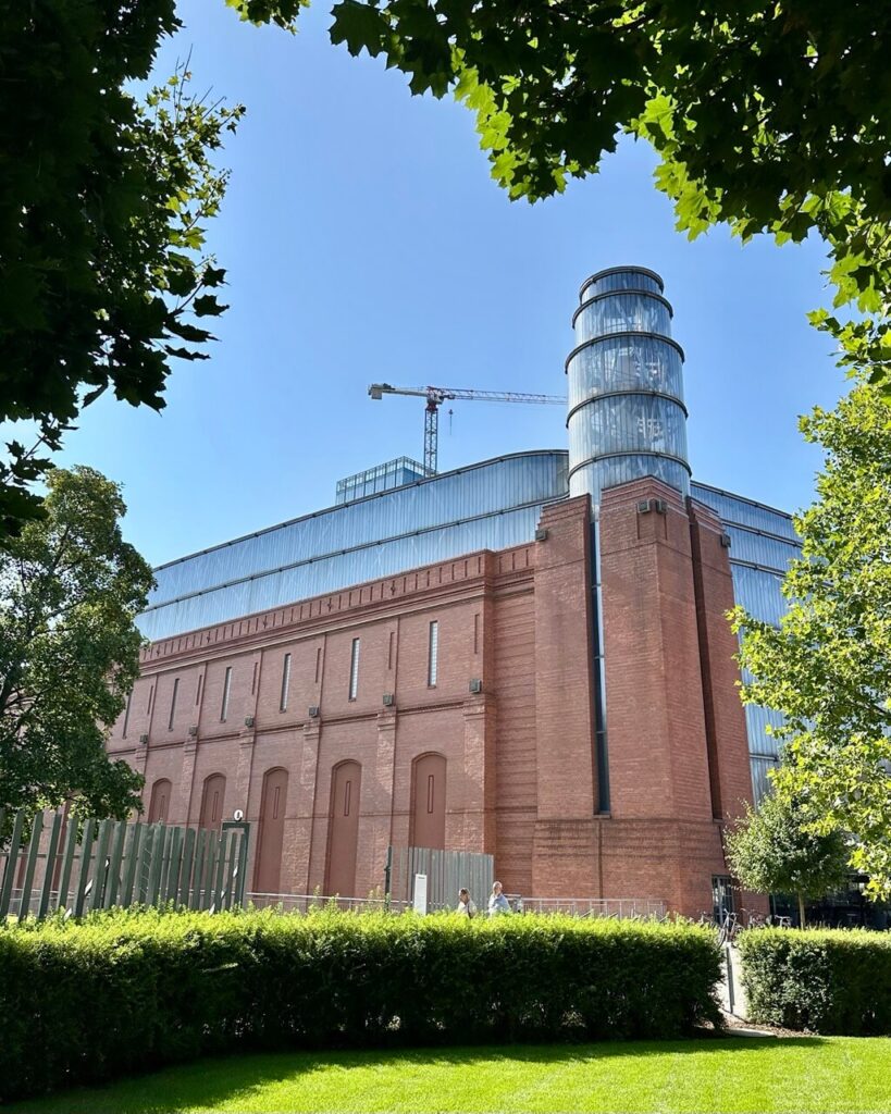 Industrial-style building with red brick base and glass cylinder tower, framed by leafy trees and blue sky.