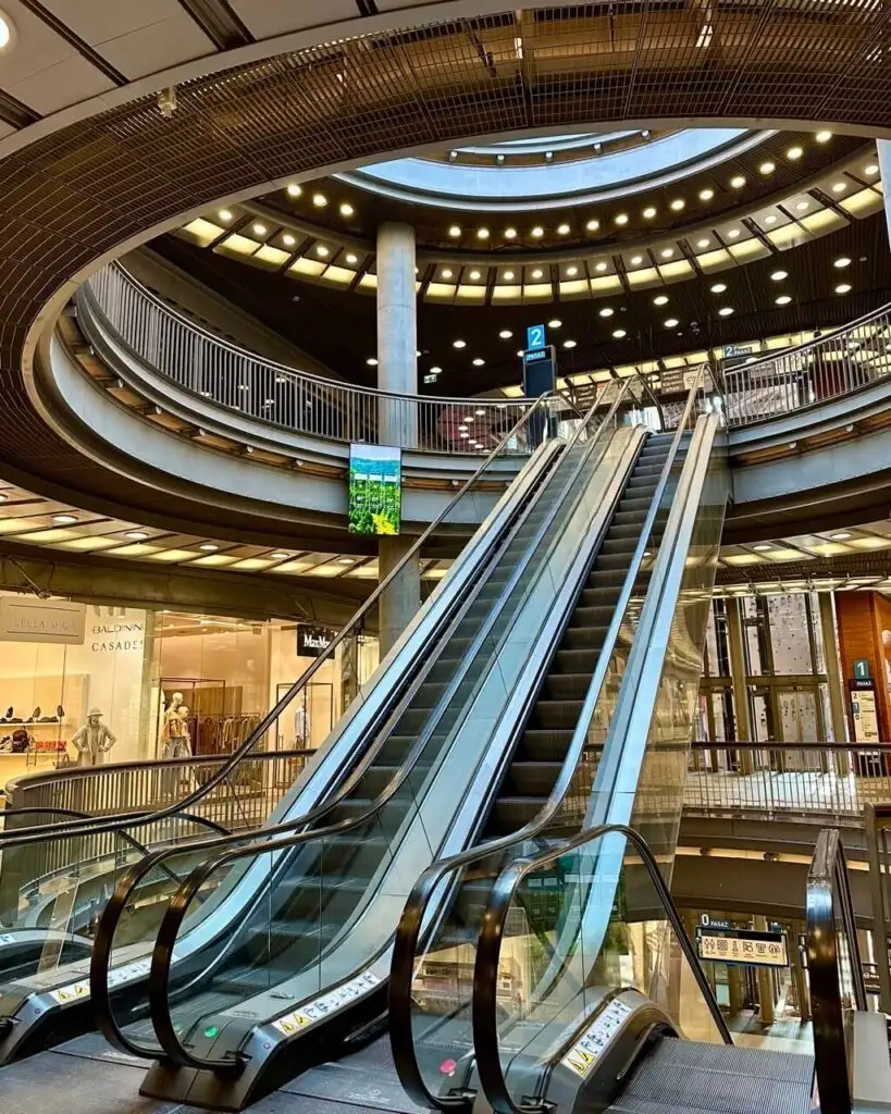 Curved escalators rise through a circular, multi-story shopping center with glowing overhead lights.