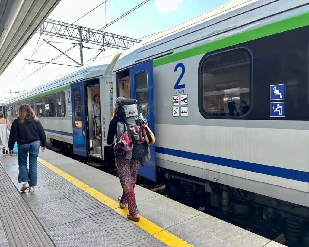 Woman with a large hiking backpack boards a blue and silver Polish train at a station platform.
