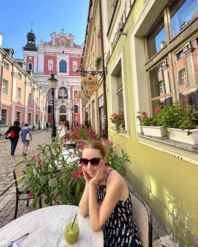 Woman in sunglasses smiles at an outdoor café table with a green drink, set on a flower-lined cobblestone street.