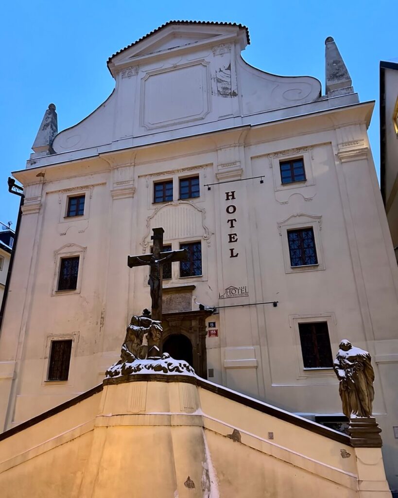 Historic Prague hotel building with a baroque style white facade and a large crucifix statue in front, located in a quiet Prague neighborhood near the city center.