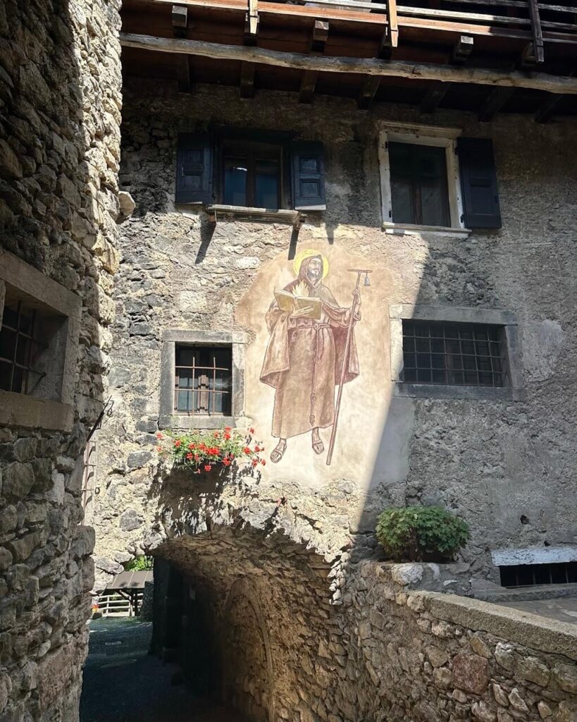 Historic stone courtyard in Canale di Tenno with a saint mural above an arched passage and red flowers in a window box.