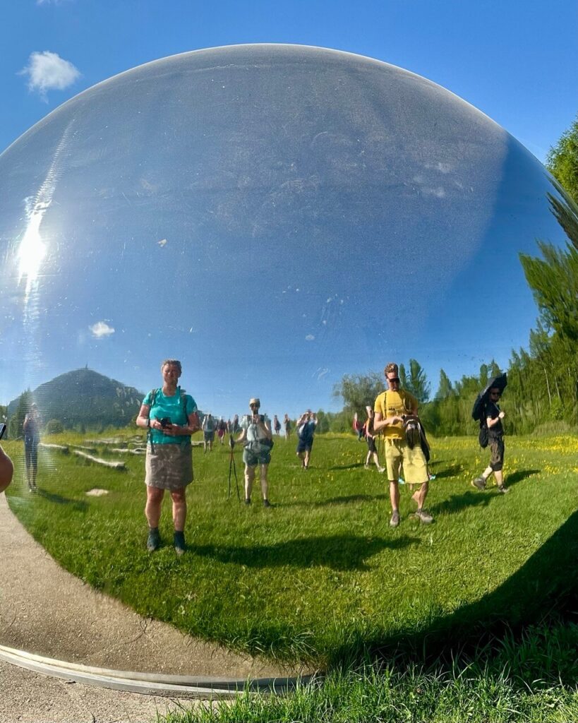 Reflective dome at the astronomical observatory near Monte Bondone shows distorted reflections of hikers. Grass. Trees. And blue sky.