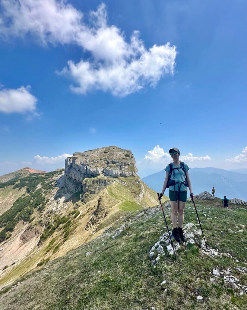 Hiker with trekking poles stands on a grassy ridge between Monte Bondone and Dos d&rsquo;Abramo with a dramatic rocky peak behind her.