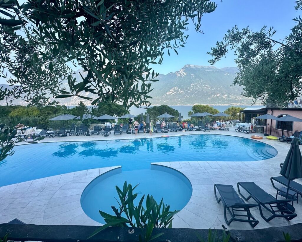 Resort pool overlooking Lake Garda Italy with mountains across the water and olive branches framing the view. People relax on sun loungers around the pool after a day outdoors.