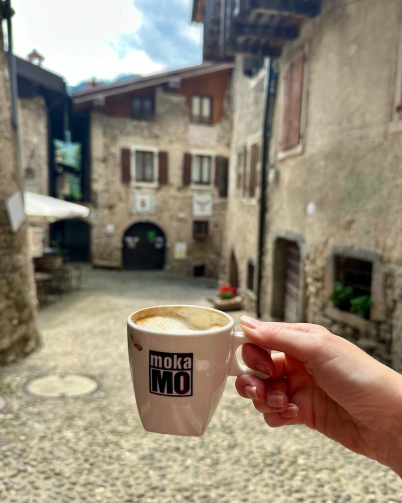 A hand holds a coffee cup in a cobblestone square surrounded by old stone buildings in Canale di Tenno. Text reads &ldquo;mokaMO&rdquo;.