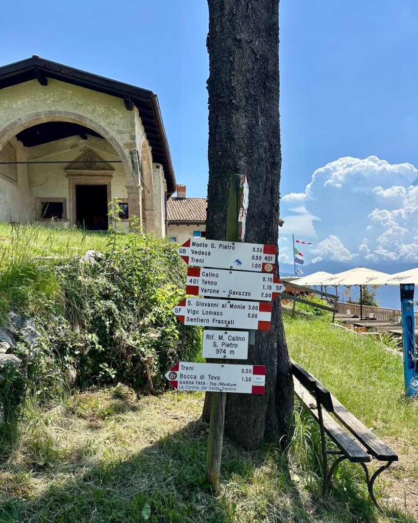 Trail signs at Rifugio Monte Calino San Pietro point toward hiking routes above Lake Garda Italy beside a small chapel and terrace. Text reads &ldquo;Monte S. Pietro 0.20. Vedese 0.50. Treni 1.00. Calino 0.15. Tenno 1.15. Varone Gavazzo 2.00. S. Giovanni al Monte 3.00. Vigo Lomaso 5.00. Sentiero Frassati. Rif. M. Calino S. Pietro 974 m. Treni 0.50. Bocca di Tovo 1.30. GARDA TREK Top Medium La Corona del Garda Trentino&rdquo;.