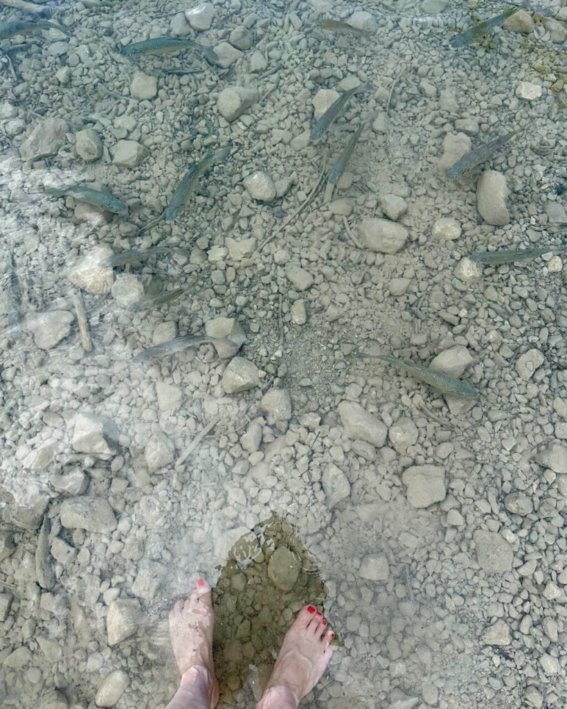 Bare feet stand in clear shallow water at Lago di Tenno while small fish swim over pale stones.