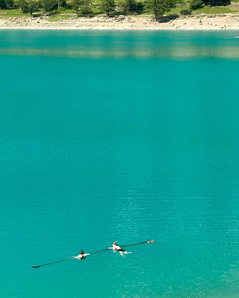 Two swimmers cross turquoise Lago di Tenno with a small dog swimming beside them and a pale beach across the water.