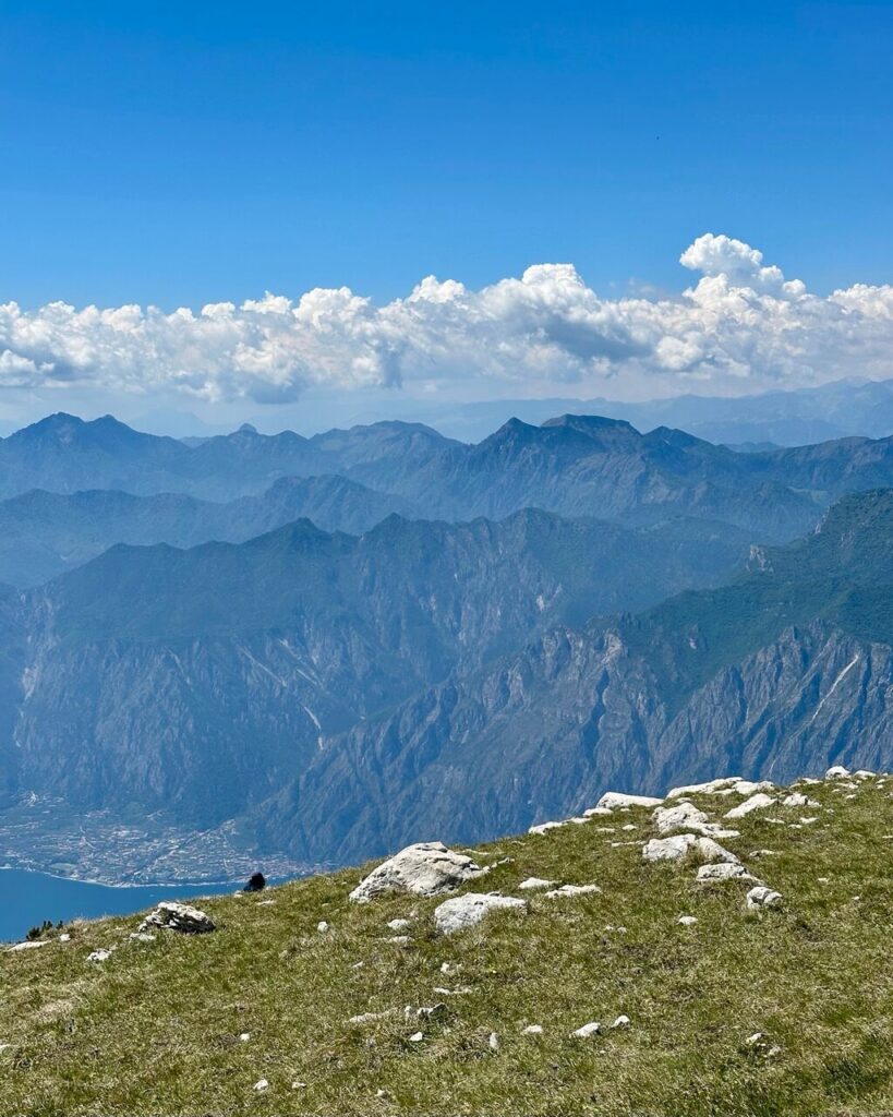 High viewpoint over Lake Garda Italy with a grassy rocky slope in the foreground and layered blue mountains under a long row of bright white clouds.