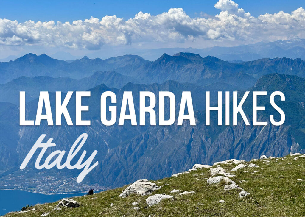 Panoramic mountain view above Lake Garda Italy with a grassy rocky foreground and hazy blue peaks stretching under puffy white clouds. Text reads "LAKE GARDA HIKES" and "Italy".