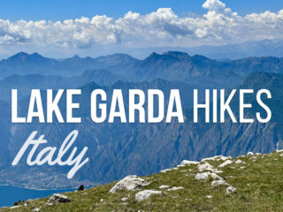 Panoramic mountain view above Lake Garda Italy with a grassy rocky foreground and hazy blue peaks stretching under puffy white clouds. Text reads 