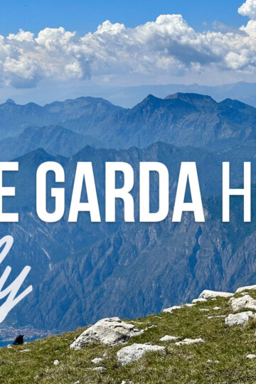 Panoramic mountain view above Lake Garda Italy with a grassy rocky foreground and hazy blue peaks stretching under puffy white clouds. Text reads "LAKE GARDA HIKES" and "Italy".