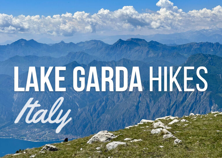 Panoramic mountain view above Lake Garda Italy with a grassy rocky foreground and hazy blue peaks stretching under puffy white clouds. Text reads "LAKE GARDA HIKES" and "Italy".