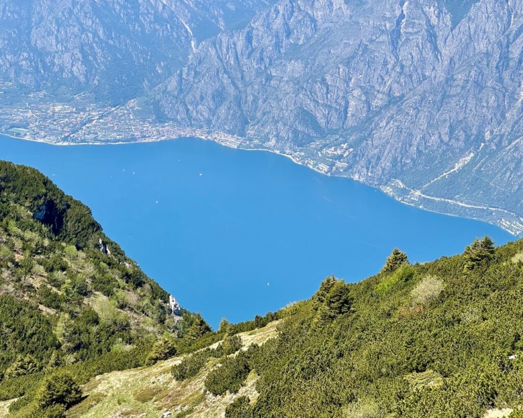 Steep green hillside drops toward the bright blue water of Lake Garda with lakeside towns tucked below rocky mountain cliffs. This high viewpoint shows the dramatic scenery along Lake Garda hikes.