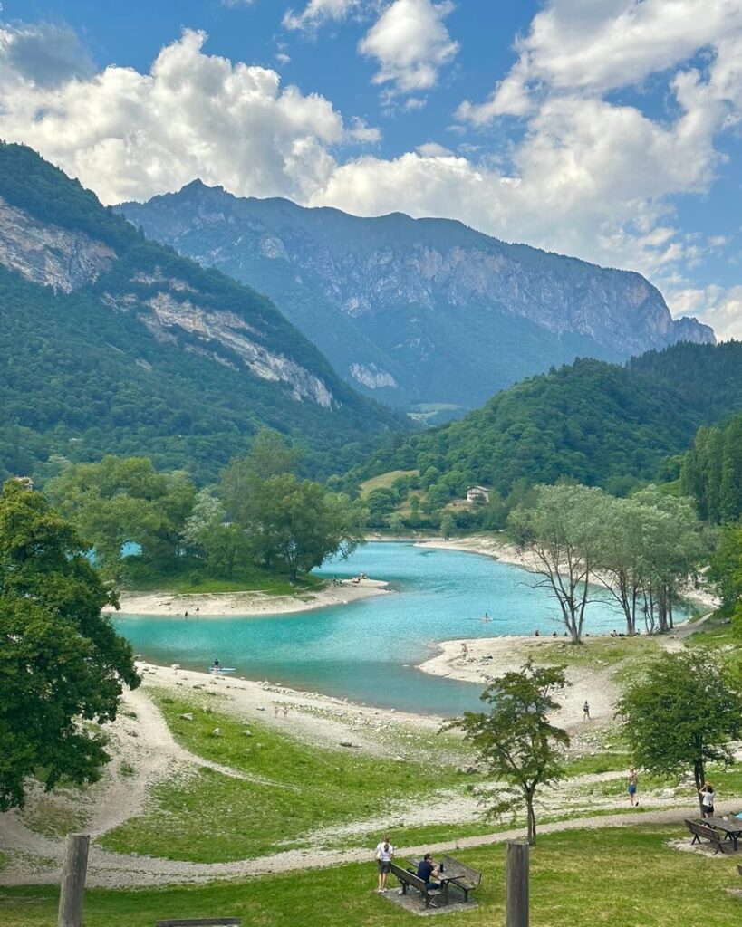 Lago di Tenno sits turquoise between forested hills and rocky mountains with small beaches and people scattered around the shore.