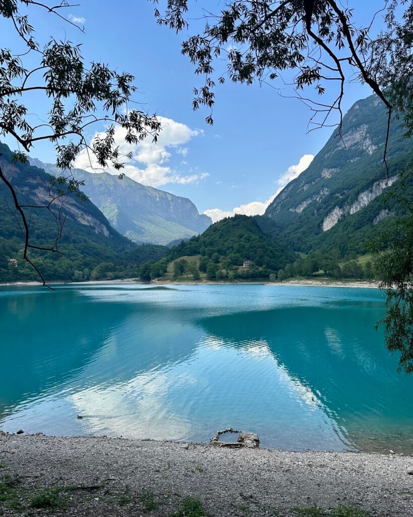 Turquoise Lago di Tenno reflects the surrounding cliffs and clouds beneath overhanging tree branches and a pebble shore.