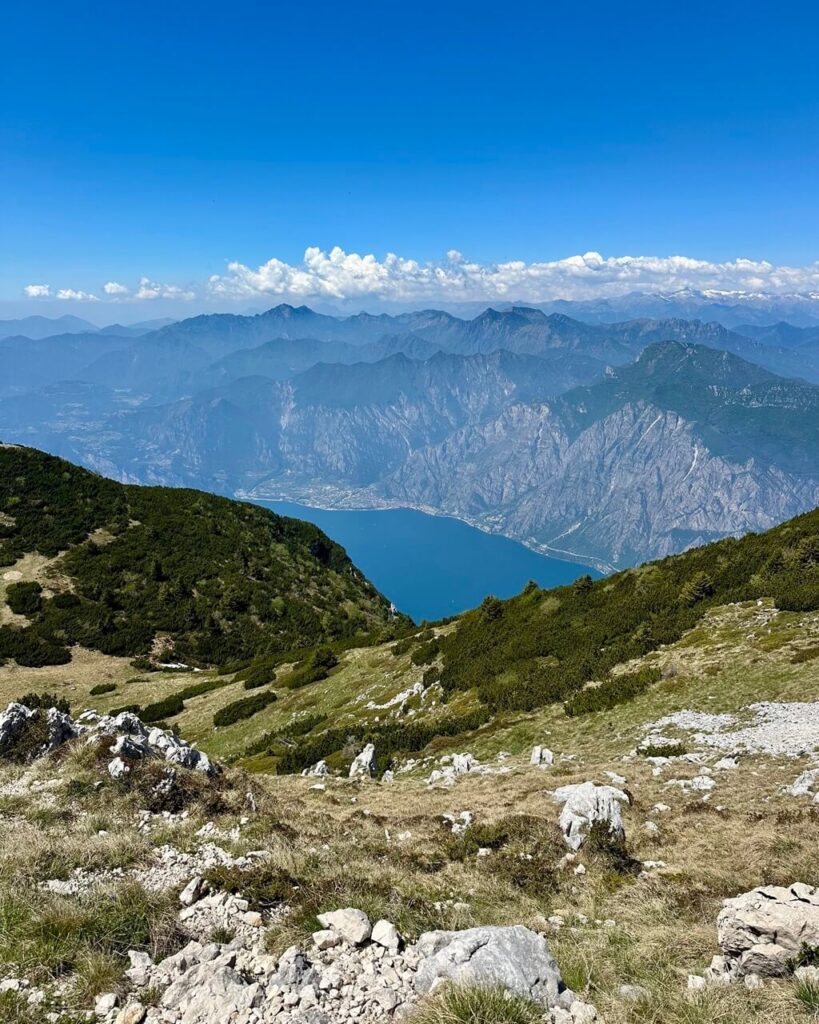 Broad view from Monte Altissimo di Nago with pale rocks. Green slopes. Blue Lake Garda water far below. Hazy mountain ridges across the horizon.