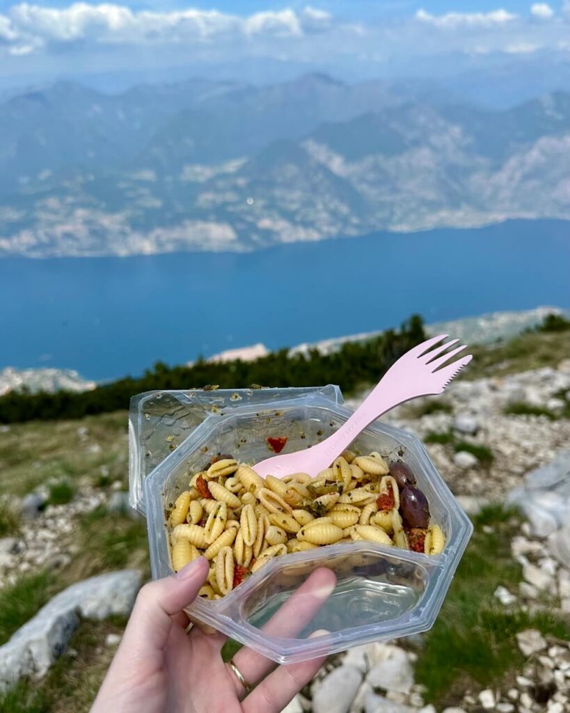 A hand holds pasta salad with a pink fork on a rocky hiking trail above Lago di Garda. The blue lake and mountain shoreline blur in the background.