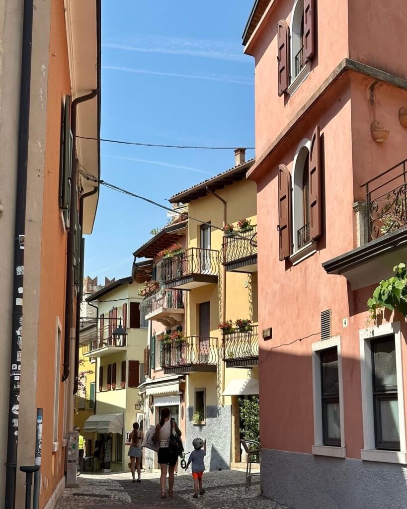 Narrow cobblestone street in a Lake Garda Italy town with pastel buildings. Wooden shutters. Flower boxes. Balconies. People walking downhill in the sun.