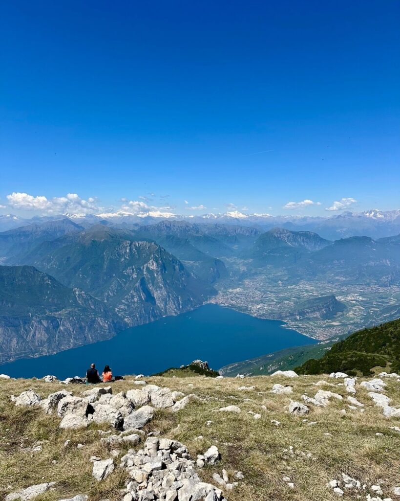 Two hikers sit on a rocky grassy ridge below Monte Altissimo di Nago overlooking Lake Garda Italy with steep cliffs. Valley towns. Snowy mountains. A clear blue sky.
