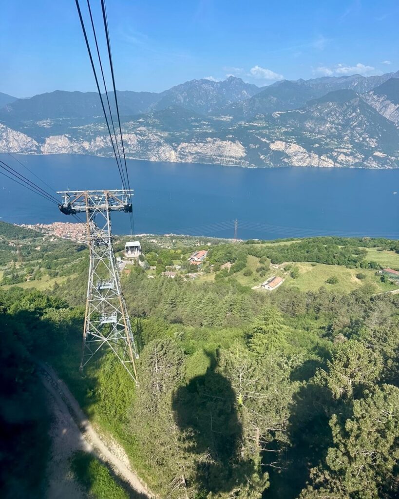 Cable car lines descend toward Lake Garda with green forest below and mountains across the water. The view connects the lift ride with easy access to Lake Garda hiking.