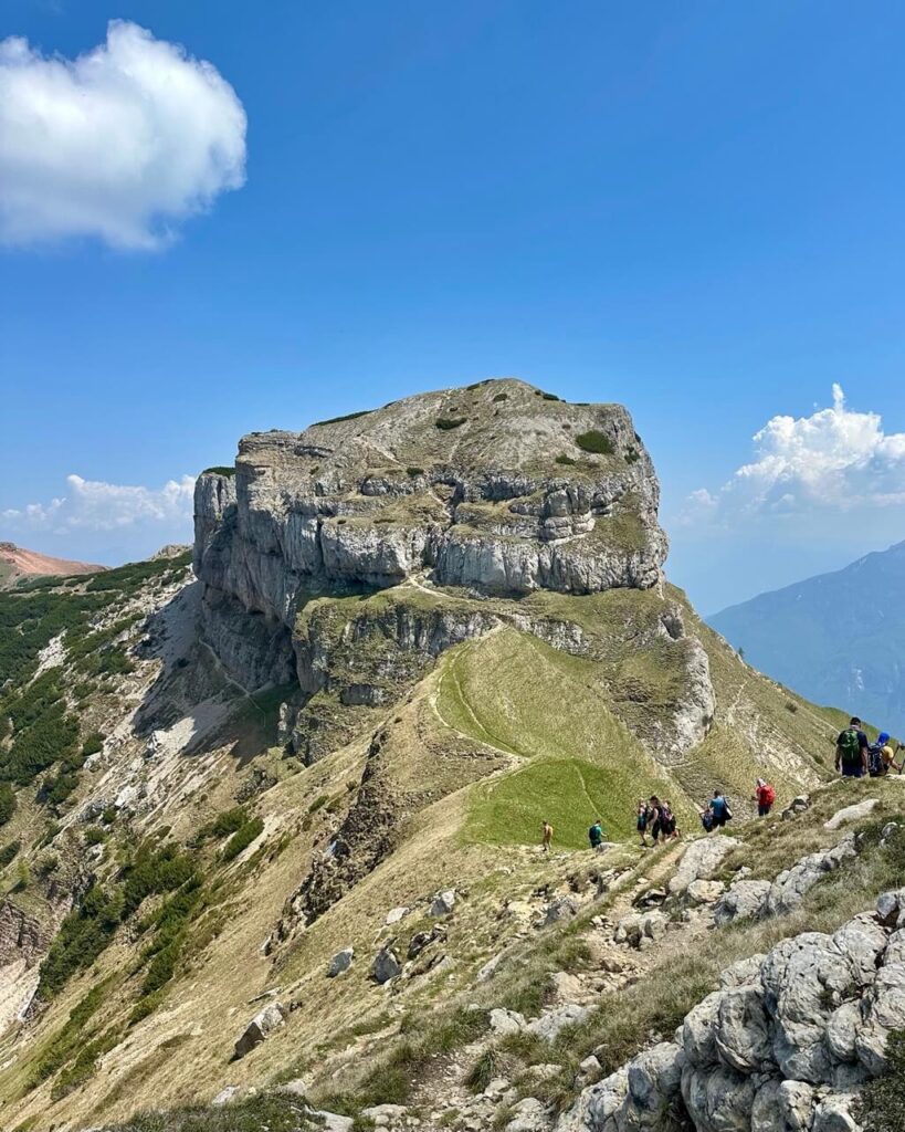 Rocky trail approaches the steep face of Dos d&rsquo;Abramo with hikers walking toward the limestone peak under a clear blue sky.