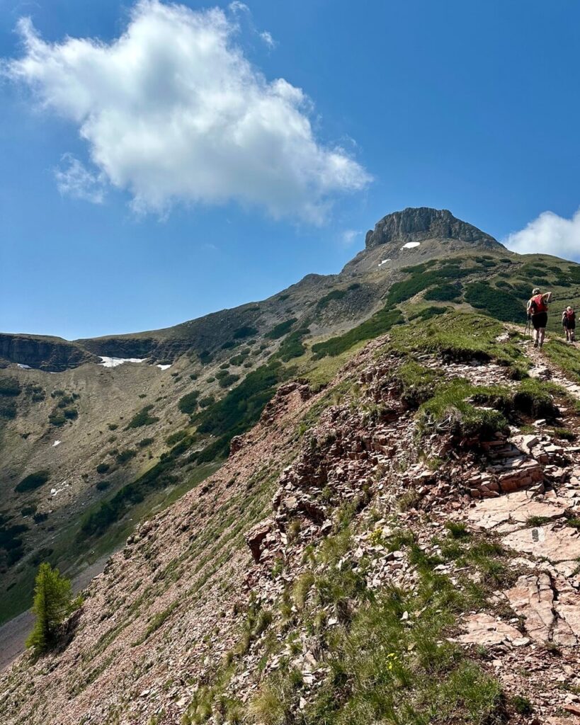 Trail climbs along a rocky red and green slope toward Monte Bondone Cornetto with small snow patches below the summit.