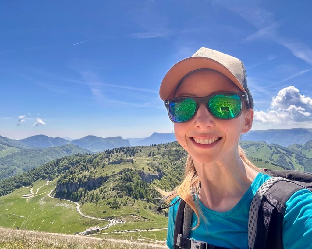 Smiling hiker wearing sunglasses and a backpack takes a selfie on Monte Altissimo di Nago with green ridges and winding mountain roads behind her.
